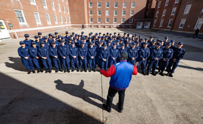 Group of uniformed personnel standing in formation outdoors in front of brick buildings, with an instructor or leader addressing them.
