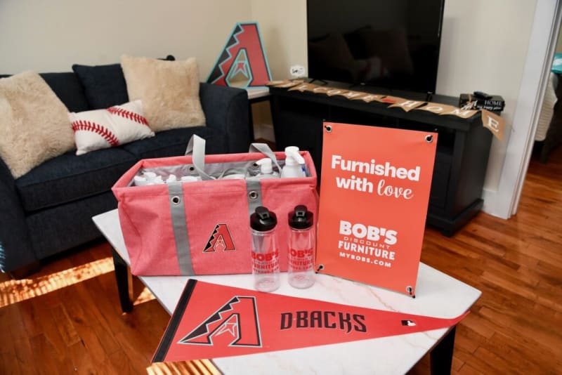 Red and gray fabric tote bag with Arizona Diamondbacks logo, two clear water bottles, and a red pennant on a white table. Next to a red sign reading: "Furnished with love. BOB'S Discount Furniture. MYBOBS.COM." Background includes a dark blue couch with baseball-themed pillows and a TV stand with a "Welcome Home" banner.