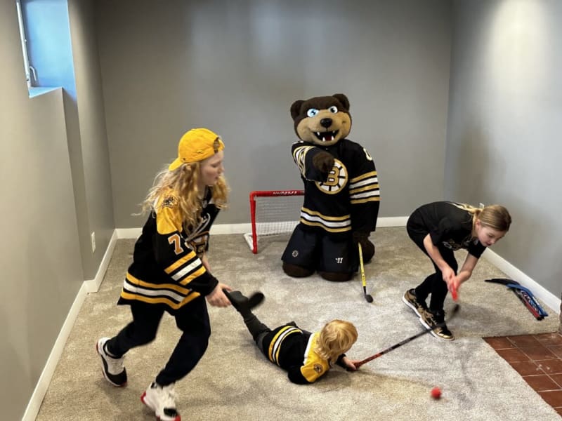 Children playing indoor floor hockey on carpeted room with a Boston Bruins mascot and mini goal, wearing black and yellow hockey jerseys.