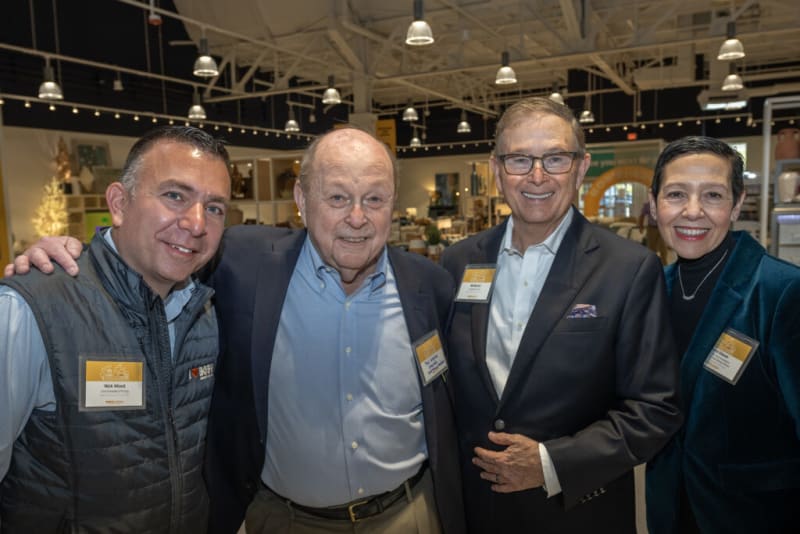 Four men wearing business casual attire and event name badges stand together in a well-lit indoor venue with high ceilings and decorative lighting.