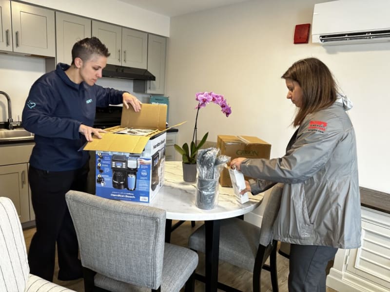 Two people unpack a Hamilton Beach coffee maker box on a round white marble table in a modern kitchen with gray cabinets.