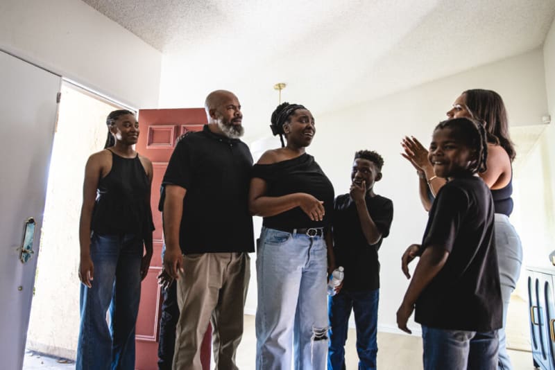 A group of diverse people standing indoors near an open door, engaged in conversation, wearing casual black tops and jeans.