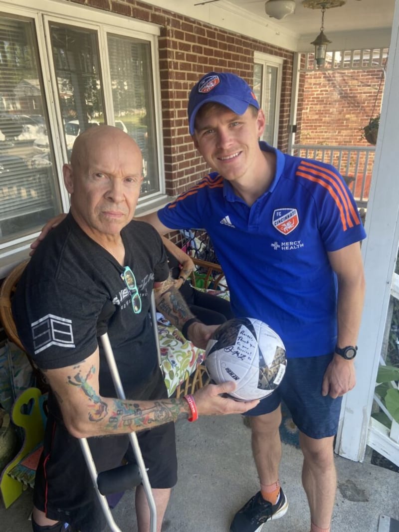 Two men on a porch, one wearing a blue FC Cincinnati polo and cap holding a signed soccer ball, the other with tattoos and crutches.