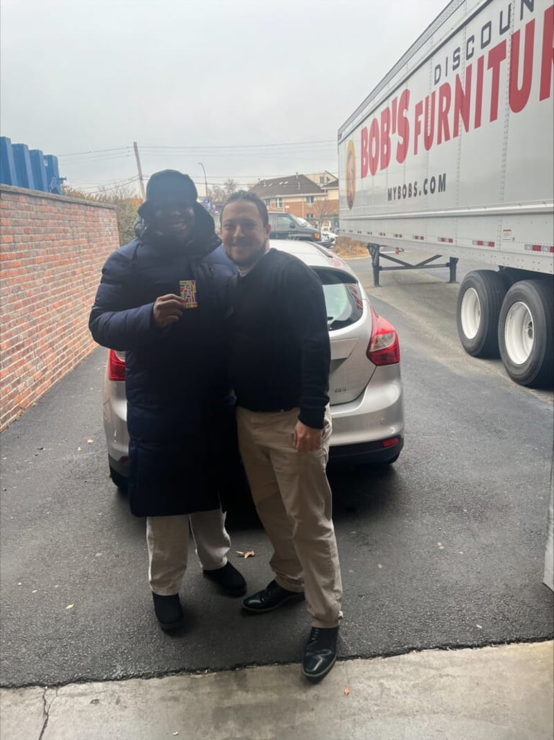 Two men standing in front of a silver car parked near a brick wall and a large truck with "Bob's Discount Furniture" branding on the side. One man wears a long dark puffer coat and light pants, holding a colorful card. The other man wears a black sweater, beige pants, and black shoes. The setting appears to be an outdoor parking or loading area.