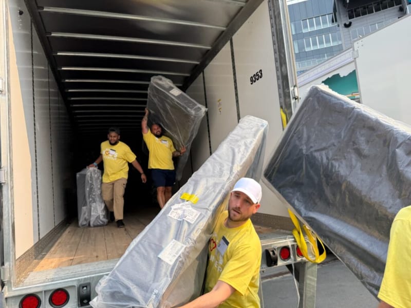 Workers wearing yellow shirts are unloading large, plastic-wrapped mattresses from a truck in an urban setting, demonstrating mattress delivery and handling.