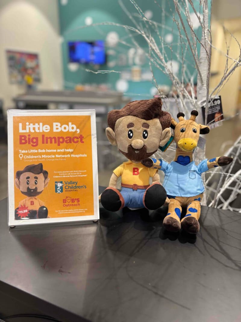 Sign promoting "Little Bob, Big Impact" plush toy fundraiser for Children's Miracle Network Hospitals, featuring a brown-haired plush figure in yellow shirt and blue pants, next to a giraffe plush in a blue shirt, priced at $11.99.