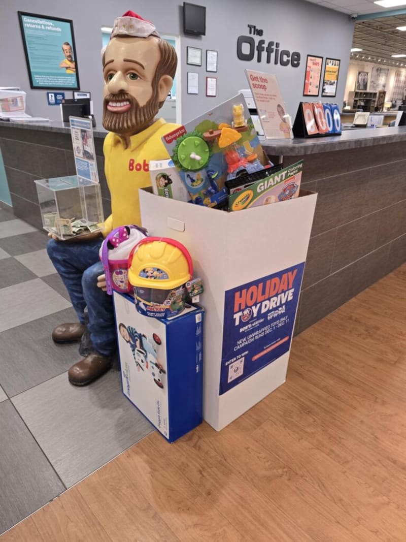 Life-sized Bob figure wearing a red Santa hat and yellow shirt holding a donation box next to a large white bin labeled "Holiday Toy Drive" filled with Fisher-Price toys, Crayola Giant coloring pack, and other children's gifts in an office lobby with "The Office" sign on the wall.