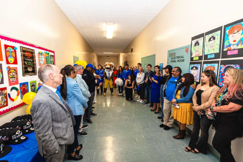 A diverse group of people standing in a hallway decorated with colorful African mask artwork and a bulletin board titled "Black History Celebrate A Rich Heritage." A mascot in a sports uniform and cheerleader are at the center, indicating a school or community event. The hallway has an "Electrical Panel Room" door and a table with black hats labeled "SBCHAMPS."
