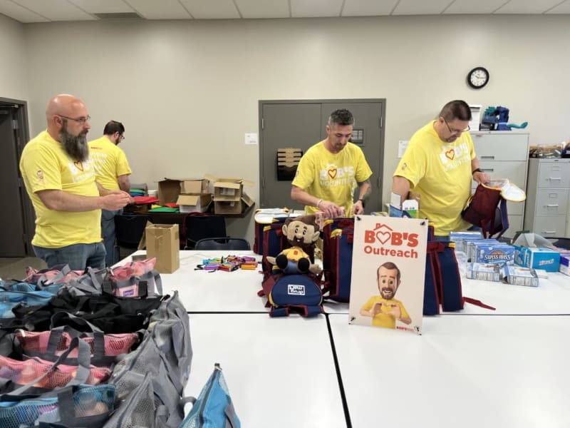 Volunteers in yellow shirts packing red and blue Bob's Outreach backpacks with supplies like Swiss Miss hot cocoa in an organized community service setting.