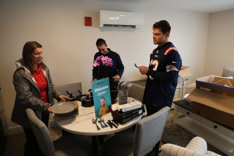 Three people unpack kitchenware around a round white marble table with gray chairs in a modern room. The table holds black pots, utensils, stacked gray plates, and a blue sign with a cartoon character and text: "Welcome home." A purple orchid is also on the table. Cardboard boxes and a white console table are visible in the background. One person wears a navy New England Patriots jersey.
