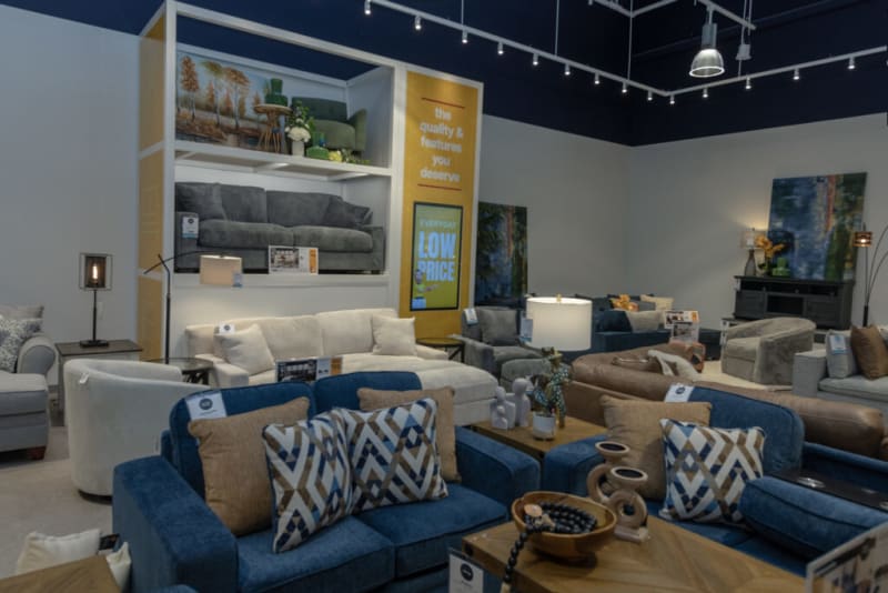 Furniture showroom display featuring various fabric sofas and armchairs in neutral tones and blue hues, with decorative pillows and modern lighting. A yellow vertical banner reads: "the quality & features you deserve" and "EVERYDAY LOW PRICE." The setting highlights contemporary living room furniture options.
