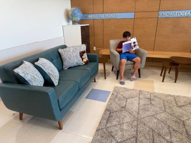Modern seating area with a teal leather sofa featuring patterned cushions and a plush toy, a beige armchair, wooden side tables, a globe, and wall panels with the text "SHARING THE PLANET" and "KNOWLEDGE."