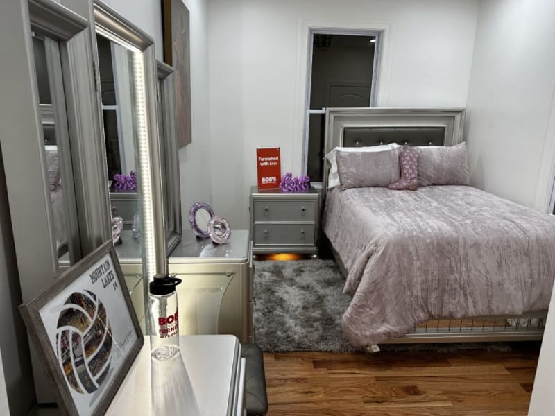 Modern bedroom setup with a silver upholstered bed featuring mauve velvet bedding, matching pillows, a nightstand with drawers, and decorative purple glass accents. The room includes a gray shag rug, wooden flooring, a large mirror with LED lighting, and a framed "Mountain Lakes" volleyball poster. The nightstand has a red sign reading "Furnished with Ease BOB'S."