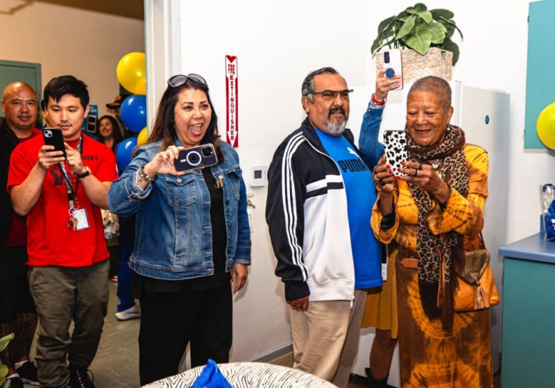Group of people holding smartphones, some taking photos or videos, standing indoors near balloons and a fire extinguisher sign.