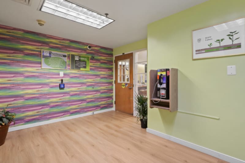 A colorful hallway with a wooden door, a mounted payphone, and two framed signs on a multicolored wall reading "MISSION" and "VISION." The mission sign states: "Compassionate, holistic services to promote wellness and independence for individuals and families in our community." The vision sign states: "For homelessness to be eradicated in this community we serve." A framed chart titled "SEED Values" with plant growth stages is on a light green wall. The floor is light wood laminate.
