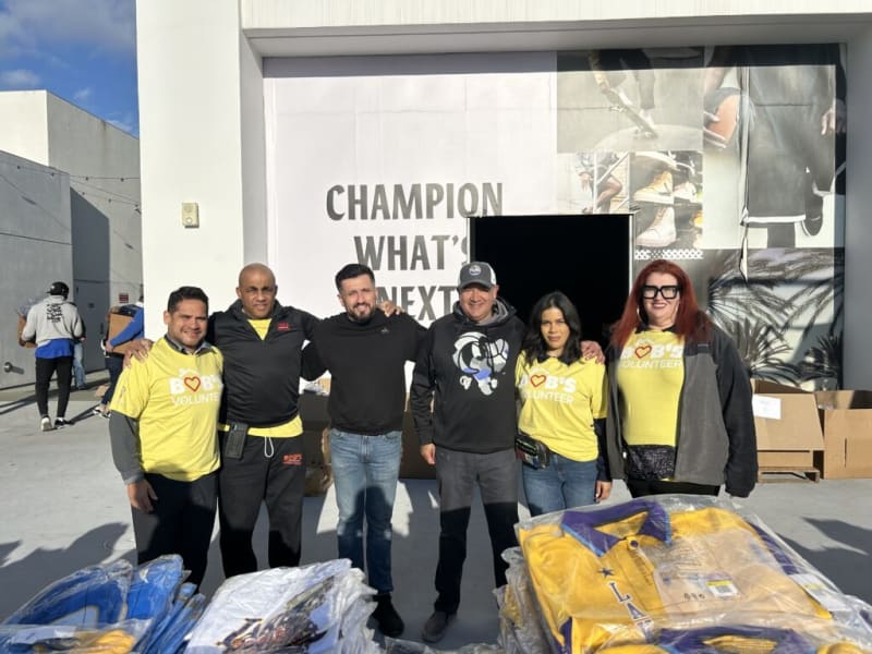 Group of six volunteers wearing yellow "B❤️B'S VOLUNTEER" shirts and casual clothing standing in front of a white building with text "CHAMPION WHAT'S NEXT" on the wall. Boxes and packaged clothing items are visible nearby, suggesting a community or charity event.