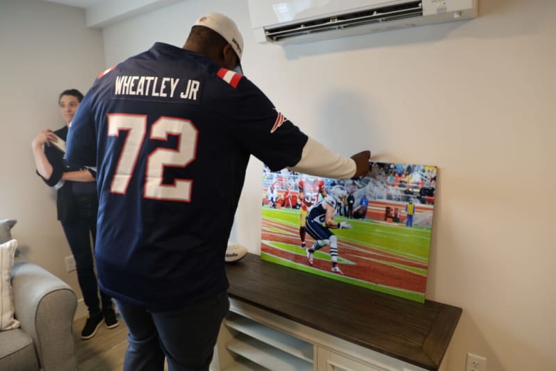 Man wearing a navy blue Wheatley Jr #72 football jersey adjusting a framed football action photo on a wooden console under a wall-mounted air conditioner.