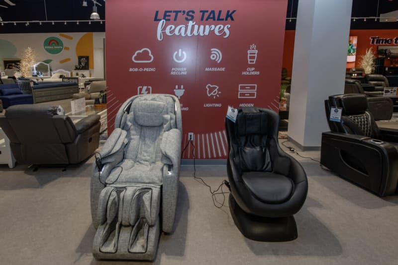 A showroom display with two massage chairs in front of a red wall featuring the text "LET'S TALK features" and icons with labels: BOB-O-PEDIC, POWER RECLINE, MASSAGE, CUP HOLDERS, LIGHTING, and HIDDEN. The chairs are upholstered in gray fabric and black leather, highlighting features like massage and power recline for comfort and relaxation.