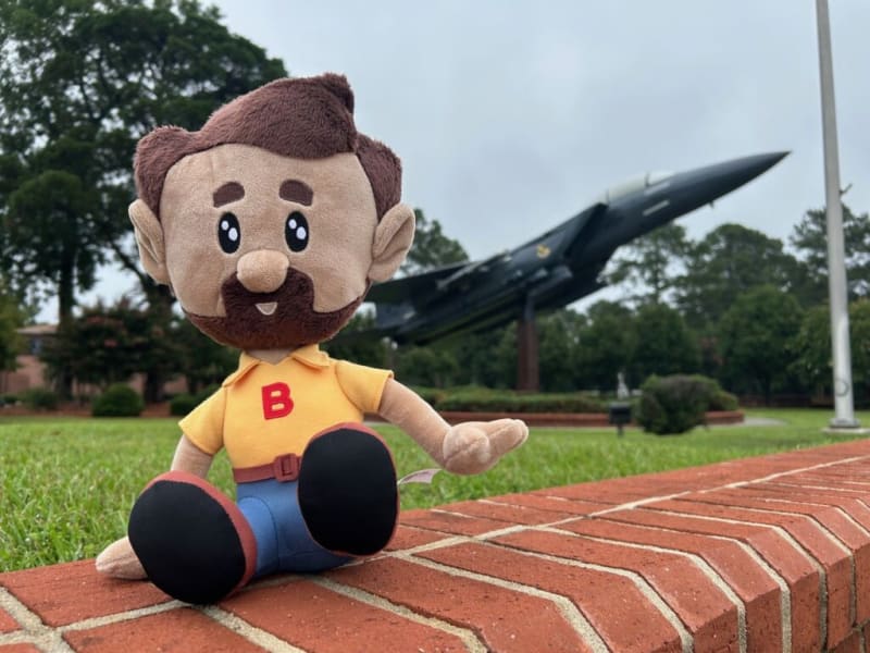 Plush toy with brown hair, beard, and mustache wearing a yellow shirt with red "B" and blue pants, sitting on a brick ledge outdoors near a jet aircraft display.