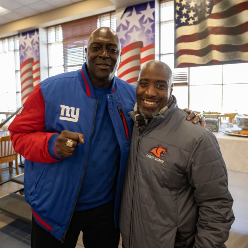 Two men standing indoors with one wearing a blue and red New York Giants jacket and the other in a gray Coast Guard jacket, with American flag decor in the background.