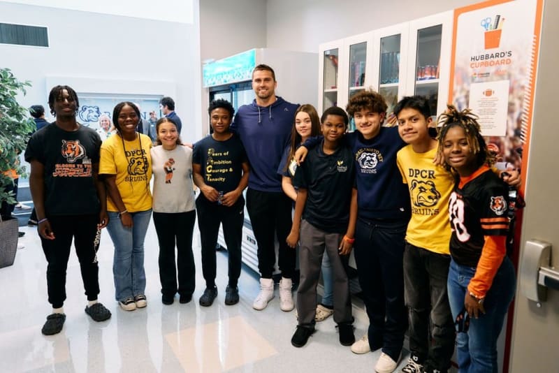 Group of diverse young people standing indoors in front of a cabinet and refrigerator, wearing casual clothing with various logos including "DPCR Bruins" and motivational text. Sign on cabinet reads "HUBBARD'S CUPBOARD" with scissors and pencil graphic.
