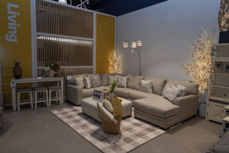 Modern beige sectional sofa with floral and solid cushions, white ottoman, plaid area rug, and decorative lighting in a living room display.