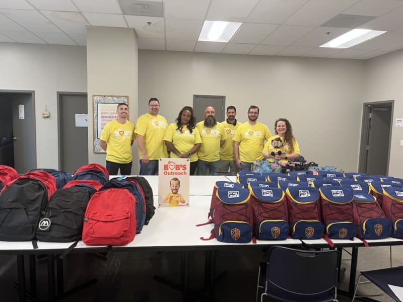 Group of volunteers wearing yellow "Bob's Outreach" t-shirts standing behind tables filled with organized red, black, and blue backpacks, promoting community outreach and support.