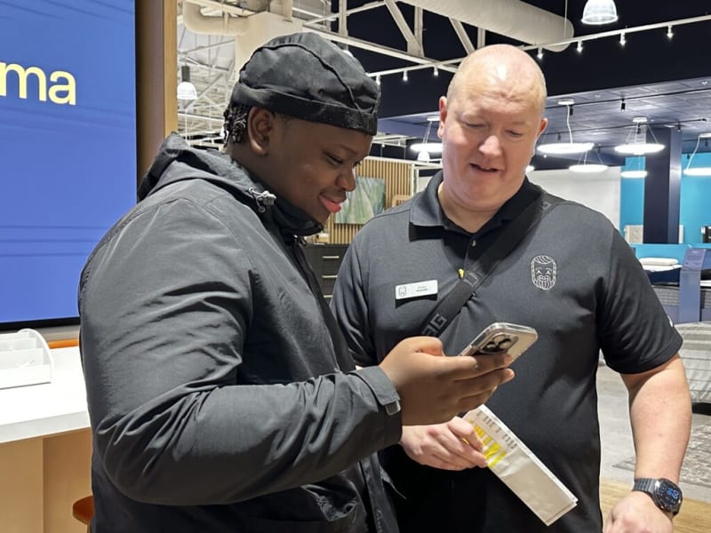 Two men in a modern retail store setting, one wearing a black jacket and cap holding a smartphone, the other in a black polo with a name tag and logo, discussing product information. The store features bright lighting, exposed ceiling pipes, and a large blue sign with partial text "ma."