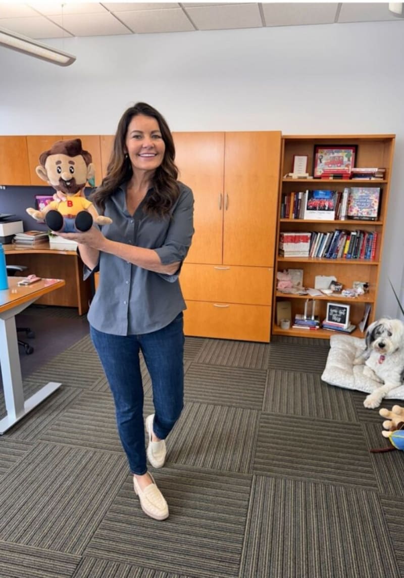 Woman standing in an office holding a plush doll with brown hair, beard, and yellow shirt with a red "B", wearing a gray button-up shirt, blue jeans, and white slip-on shoes, with wooden cabinets and a bookshelf in the background, and a white dog lying on a cushion to the right.