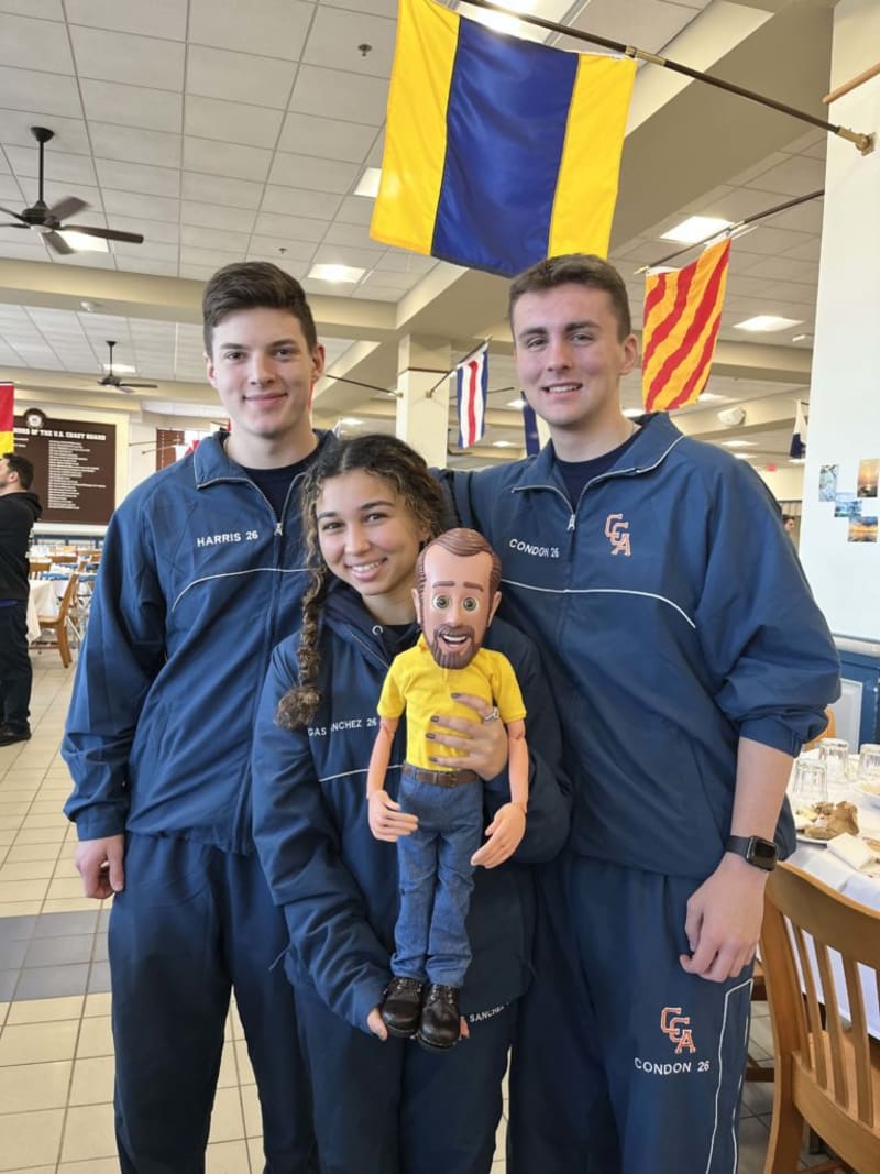 Three people wearing matching navy blue athletic jackets and pants with "GA" and names "Harris 26," "Sanchez 26," and "Condon 26" embroidered, standing indoors under nautical signal flags, one holding a doll dressed in a yellow shirt and blue jeans.