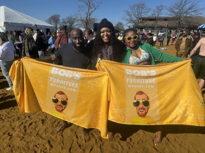 Three people stand barefoot on sandy ground holding large yellow towels with white text reading "BOB'S DISCOUNT FURNITURE MYBOBS.COM" at an outdoor event with tents and trees in the background.