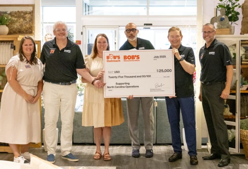 Six people stand indoors holding a large check from Bob's Discount Furniture Outreach dated July 2025, made out to USO for $25,000 supporting North Carolina operations.