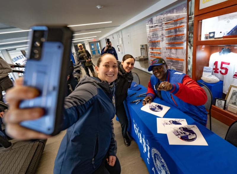 A person takes a selfie with a sports figure signing autographs at a United States Coast Guard event, with memorabilia and jerseys displayed.