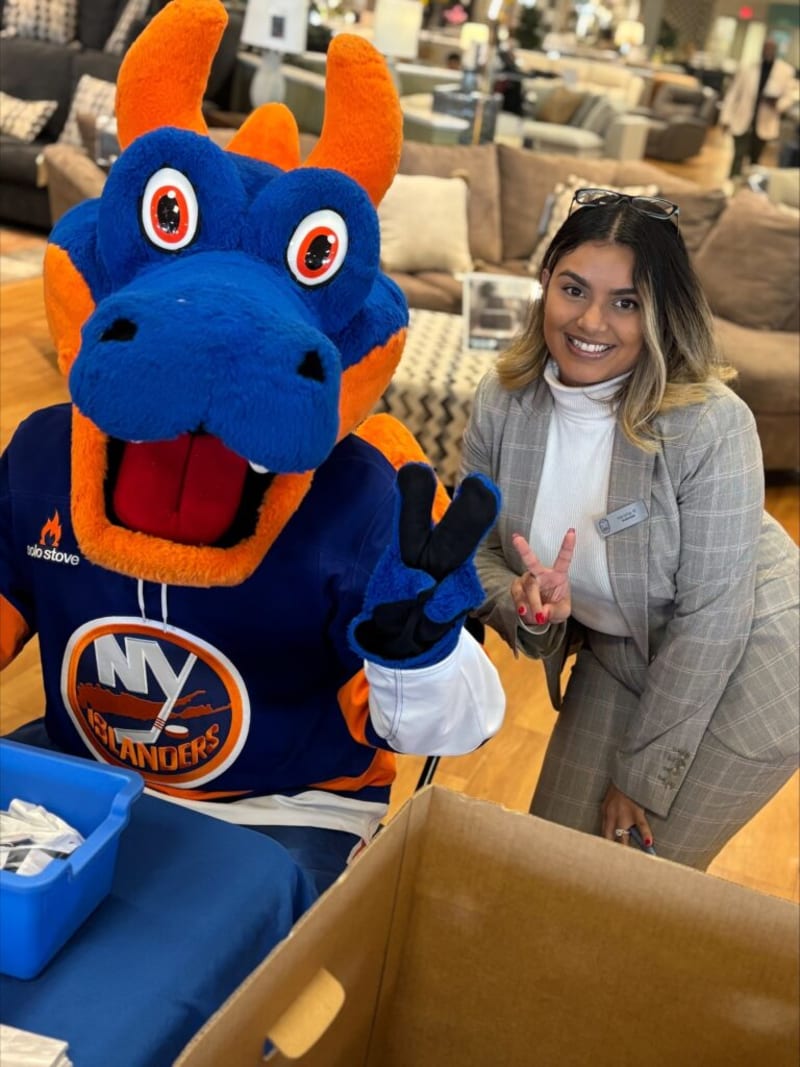 Person in a gray plaid suit and white turtleneck posing with a blue and orange NY Islanders mascot making a peace sign indoors.