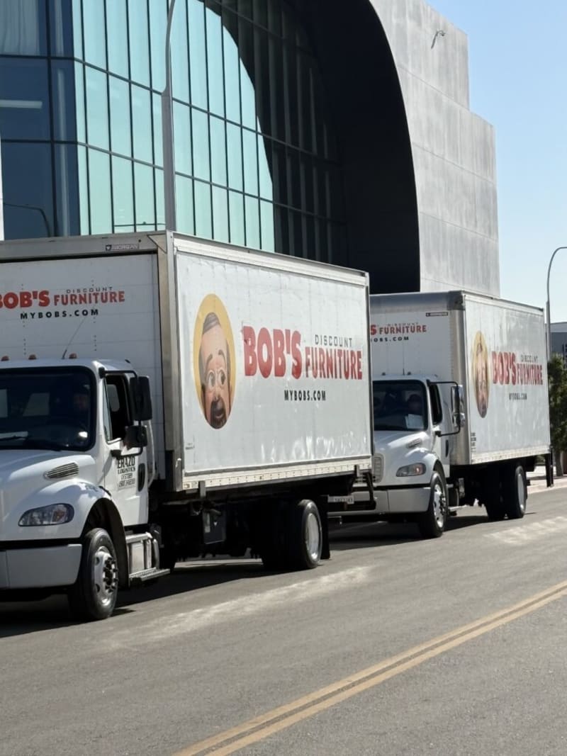 Two white delivery trucks parked on a street in front of a modern glass building. Both trucks display large side panels with the text "BOB'S DISCOUNT FURNITURE MYBOBS.COM" and cartoon faces, promoting Bob's Discount Furniture brand.