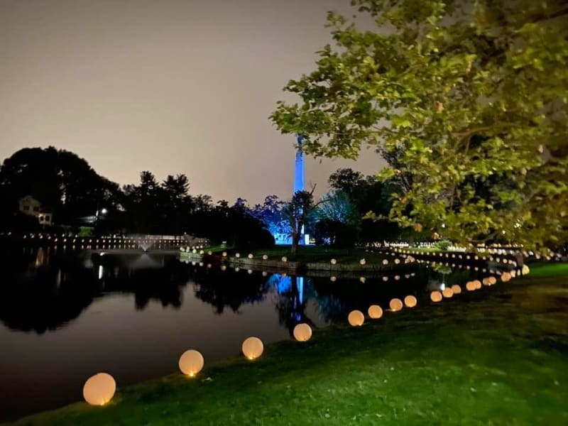 A nighttime scene of a serene pond surrounded by glowing spherical lights along the grassy shore, with a tall illuminated blue monument and trees in the background.