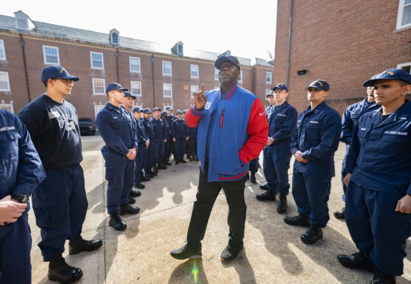 A man in a blue and red New York Giants jacket stands between two lines of U.S. Coast Guard personnel in navy uniforms outdoors near brick buildings.