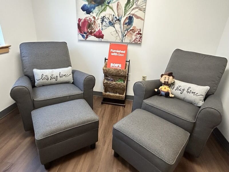 Two gray upholstered armchairs with matching ottomans, each featuring a rectangular pillow embroidered with "let's stay home." A small basket stand between the chairs holds a red sign reading "Furnished with love BOB'S." The setting suggests a cozy, inviting living room or waiting area with wood flooring and floral wall art.