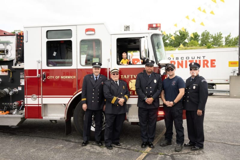 Five firefighters in uniform standing in front of a red and white fire truck labeled "CITY OF NORWICH" and "TAFTVILLE FIRE."