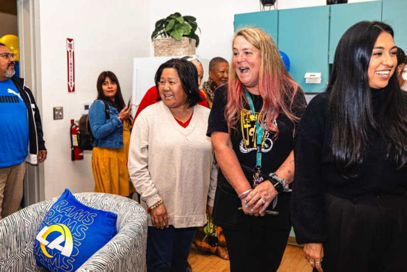 Group of diverse adults standing in a room with blue Rams-themed pillow on zebra-patterned chair, wearing casual clothing including a Puma shirt and lanyard.