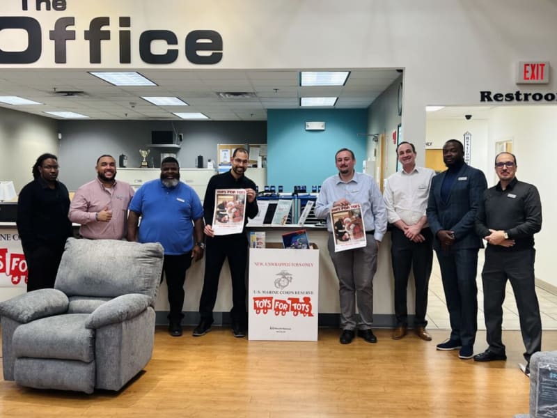 Seven men stand in an office lobby under a large sign reading "The Office," holding Toys for Tots flyers above a donation box labeled "NEW, UNWRAPPED TOYS ONLY U.S. MARINE CORPS RESERVE TOYS FOR TOTS." A gray upholstered recliner is in the foreground, and an exit sign and restroom sign are visible on the wall.