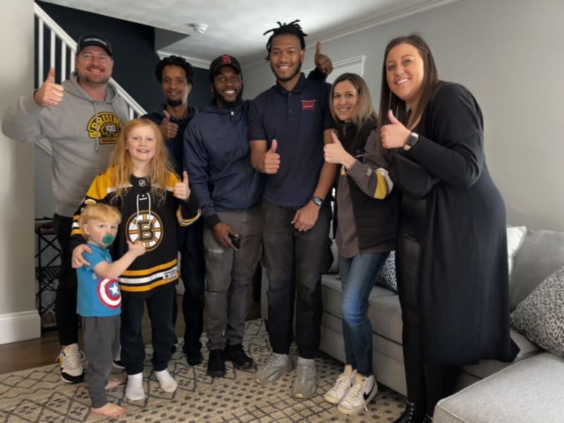 Group of diverse people, including children and adults, standing indoors on a patterned rug near a gray sofa, all giving thumbs up, with some wearing casual sports apparel including a Boston Bruins hockey jersey and Captain America t-shirt.