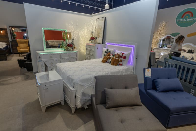 White wooden bedroom set with LED-lit mirror and headboard, featuring a bed with white quilt, decorative pillows, and gingerbread plush toys, alongside a nightstand and dresser in a showroom setting.