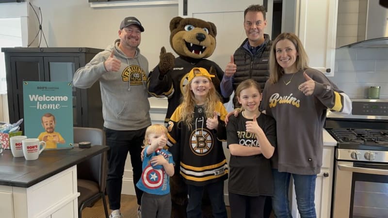 Group of people and children giving thumbs up in a kitchen, with one person in a Boston Bruins mascot costume and others wearing Bruins apparel. A teal sign on the left reads "BOB'S Furniture Welcome home."