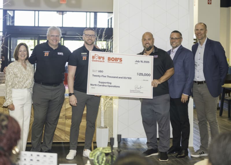 Group of six people standing indoors holding a large check from Bob's Discount Furniture for $25,000 dated July 18, 2025, supporting USO North Carolina operations.