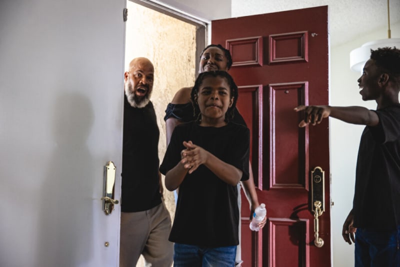 A group of people standing near an open red front door inside a home, with one person pointing and another holding a water bottle.