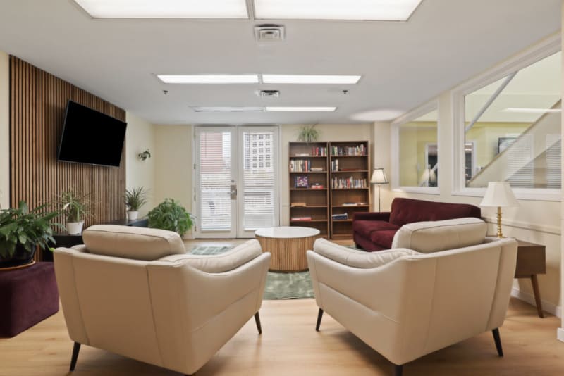 Modern office lounge with beige leather armchairs, a burgundy sofa, wooden bookshelves, plants, and a wall-mounted TV.