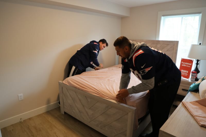 Two people wearing dark sports jerseys making a bed with a light pink fitted sheet on a wooden bed frame in a bright, modern bedroom. A nightstand with a lamp and a red sign reading "Furnished with love BOB'S DISCOUNT FURNITURE" is visible by the window.