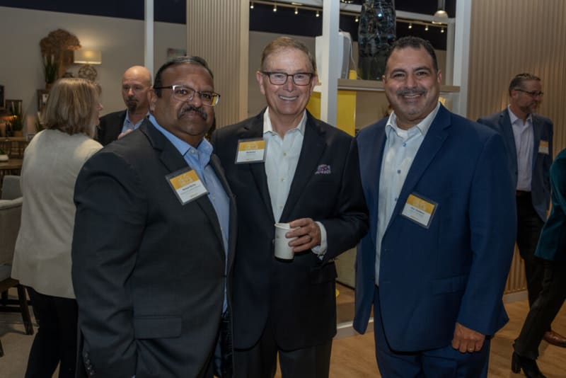 Three men in business suits with name badges at a professional networking event, one holding a white cup, indoor setting with modern decor.