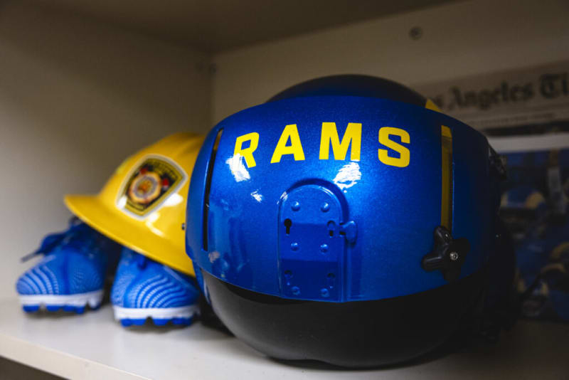 Blue and black Rams-branded helmet with yellow text "RAMS" on a shelf next to a yellow firefighter helmet and blue cleats.